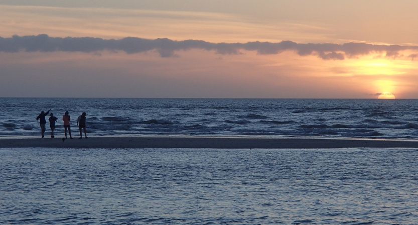 Four people stand on a small peninsula surrounded by blue water. In the background, the sun is setting on the horizon, illuminating the sky in soft shades in yellow, orange, pink and purple. 